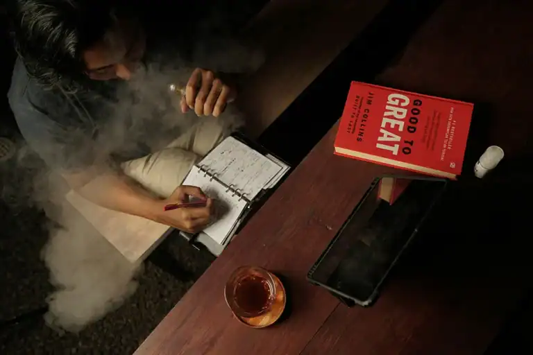 成年使用者在桌前書寫筆記並使用電子煙,桌上放有平板與書籍